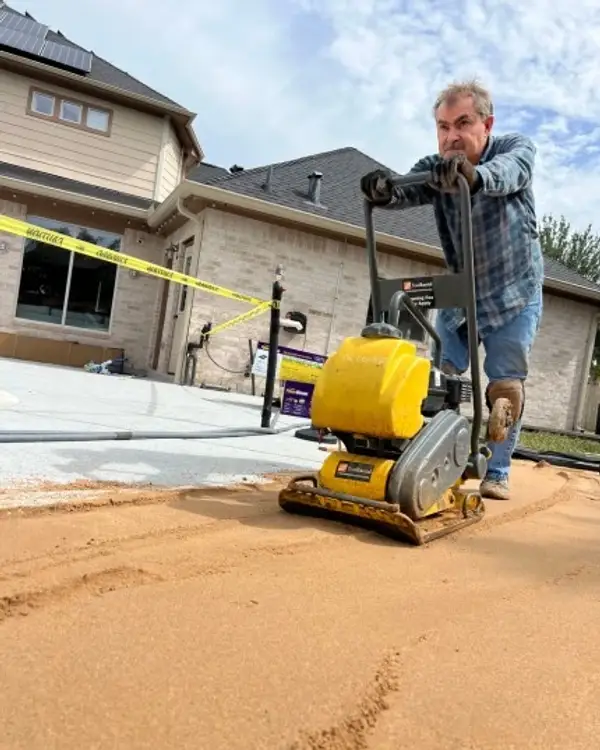 Worker running a plate compactor over sand base material in a residential side yard