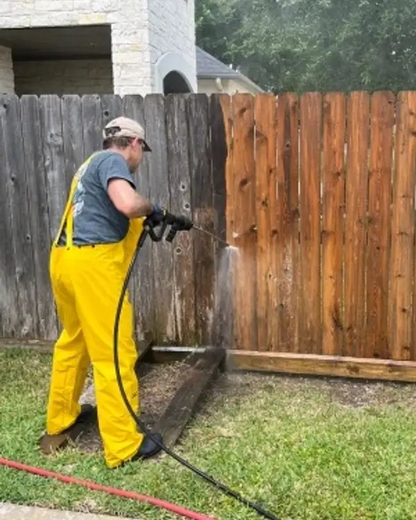 Worker pressure washing a wooden fence, with the cleaned boards visibly lighter than the rest