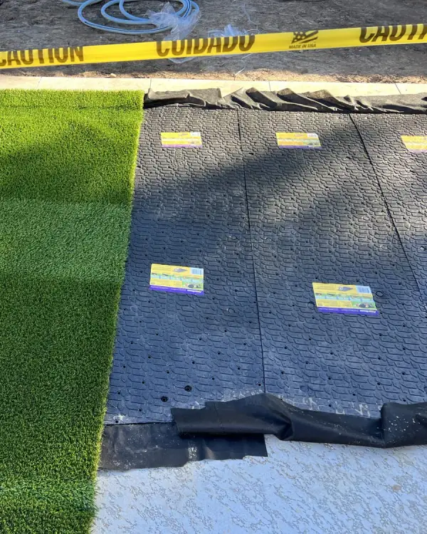 Compacted base and weed barrier laid out next to finished artificial grass, marked off with caution tape