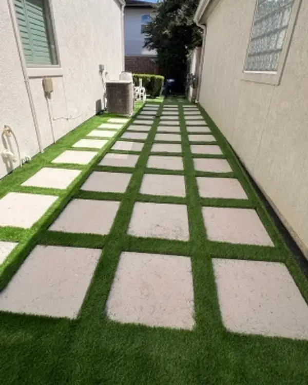 Artificial grass walkway with evenly spaced square concrete pavers between two houses