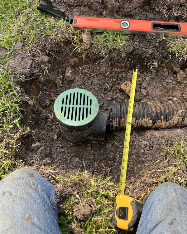 Catch basin and corrugated drain pipe set into a trench in a lawn, with a level and tape measure alongside