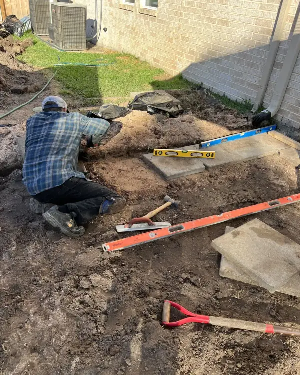 Worker digging a drainage trench beside a brick house next to the air conditioning unit